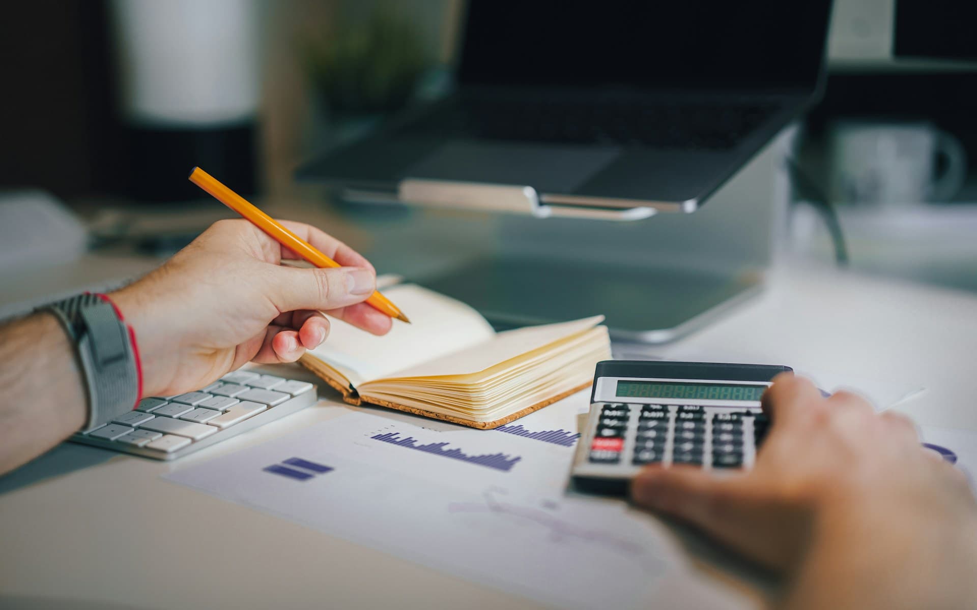 Une personne calcule un budget dans un carnet à côté d’un ordinateur